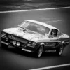 Ford Mustang Shelby GT500 Eleanor en blanco y negro en acción sobre pista de carreras, vista frontal tres cuartos con bandas deportivas y faros dobles