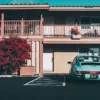 Porsche 911 SC turquesa metalizado estacionado frente a motel vintage californiano años 70, fachada beige con buganvillas rojas, balcones de madera y palmera, luz dorada ambiente retro