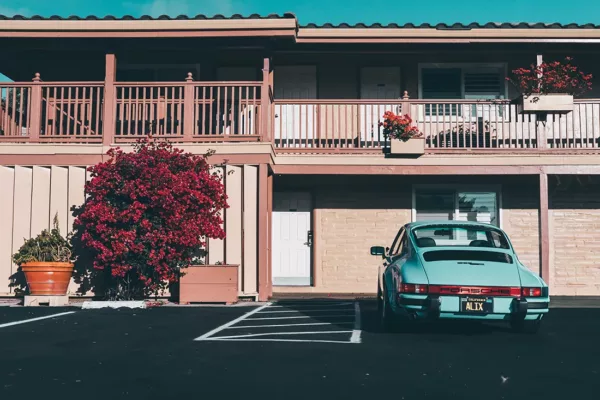 Porsche 911 SC turquesa metalizado estacionado frente a motel vintage californiano años 70, fachada beige con buganvillas rojas, balcones de madera y palmera, luz dorada ambiente retro