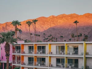Hotel The Saguaro con fachadas multicolores rosa naranja y amarilla al atardecer, palmeras del desierto y montañas San Jacinto iluminadas en tonos cálidos en Palm Springs California