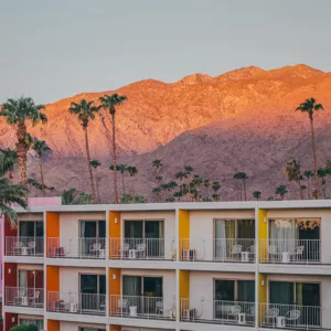 Hotel The Saguaro con fachadas multicolores rosa naranja y amarilla al atardecer, palmeras del desierto y montañas San Jacinto iluminadas en tonos cálidos en Palm Springs California