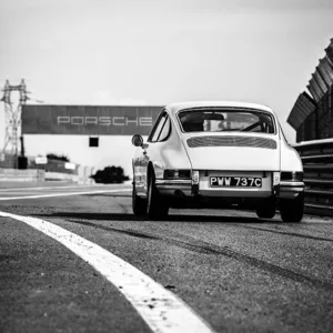 Fotografía blanco y negro de Porsche 911 clásico en pista de circuito con matrícula PWM 737C, semáforo a la izquierda, cartel Porsche al fondo y vallas de seguridad, vista trasera del vehículo en movimiento