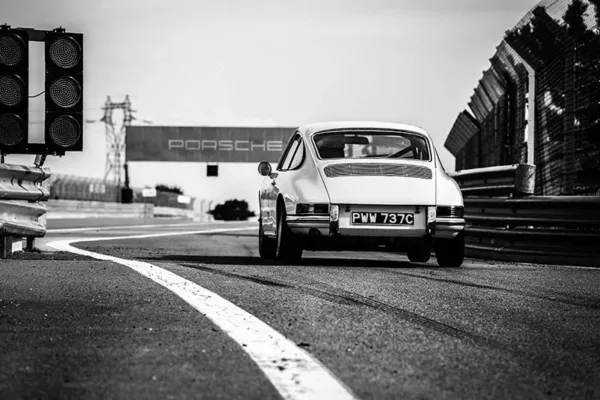 Fotografía blanco y negro de Porsche 911 clásico en pista de circuito con matrícula PWM 737C, semáforo a la izquierda, cartel Porsche al fondo y vallas de seguridad, vista trasera del vehículo en movimiento