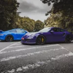 Fotografía de Porsche 991 GT3 en azul y GT3 RS en púrpura estacionados en una carretera, con detalles de los frenos amarillos y fondo de vegetación, vista lateral de ambos vehículos.