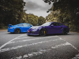 Fotografía de Porsche 991 GT3 en azul y GT3 RS en púrpura estacionados en una carretera, con detalles de los frenos amarillos y fondo de vegetación, vista lateral de ambos vehículos.