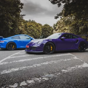 Fotografía de Porsche 991 GT3 en azul y GT3 RS en púrpura estacionados en una carretera, con detalles de los frenos amarillos y fondo de vegetación, vista lateral de ambos vehículos.
