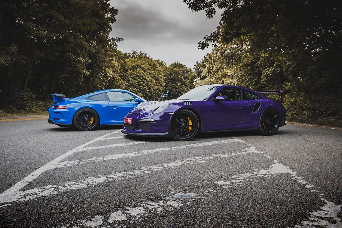 Fotografía de Porsche 991 GT3 en azul y GT3 RS en púrpura estacionados en una carretera, con detalles de los frenos amarillos y fondo de vegetación, vista lateral de ambos vehículos.