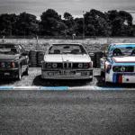 Tres BMW clásicos de competición en línea de salida Le Mans Classic en blanco y negro, BMW 2002 Turbo con bandas M tricolor, BMW Serie 3 E30 cabrio y BMW Serie 6 635CSi al fondo