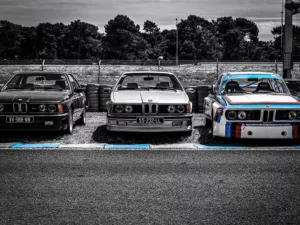 Tres BMW clásicos de competición en línea de salida Le Mans Classic en blanco y negro, BMW 2002 Turbo con bandas M tricolor, BMW Serie 3 E30 cabrio y BMW Serie 6 635CSi al fondo