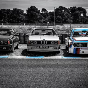 Tres BMW clásicos de competición en línea de salida Le Mans Classic en blanco y negro, BMW 2002 Turbo con bandas M tricolor, BMW Serie 3 E30 cabrio y BMW Serie 6 635CSi al fondo