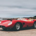Imagen 2: Fotografía de Ferrari Testarossa roja en una carretera costera, con matrícula visible, cielo nublado y paisaje marítimo al fondo, vista lateral del vehículo.