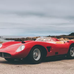 Imagen 2: Fotografía de Ferrari Testarossa roja en una carretera costera, con matrícula visible, cielo nublado y paisaje marítimo al fondo, vista lateral del vehículo.