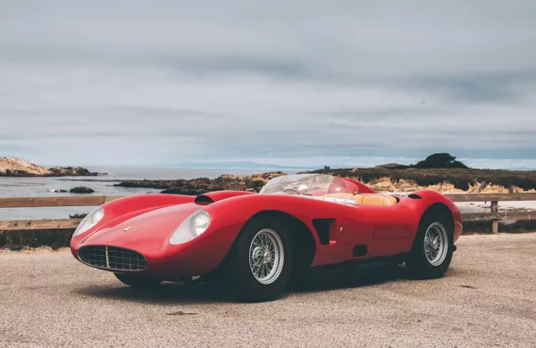 Imagen 2: Fotografía de Ferrari Testarossa roja en una carretera costera, con matrícula visible, cielo nublado y paisaje marítimo al fondo, vista lateral del vehículo.