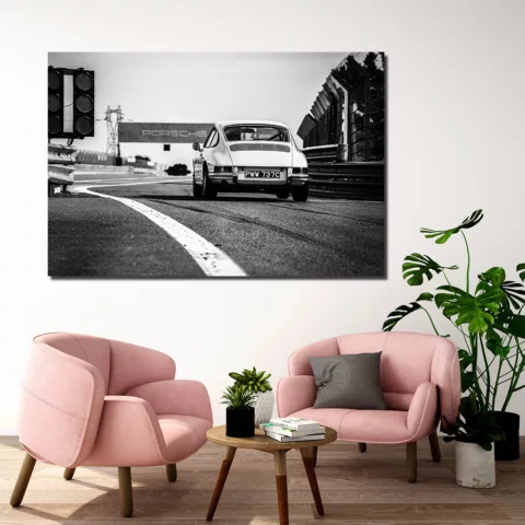 Salón moderno con cuadro grande de Porsche 911 Carrera blanco y negro en circuito colgado en pared beige, dos sillones rosas, mesa redonda de madera y plantas verdes, decoración interior contemporánea
