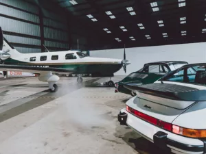 Porsche 911 Turbo blanco y 911 verde frente a un Piper Malibu en un hangar con lucernarios - cuadro fotográfico de autor