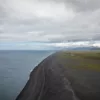 Playa de arena negra en la costa de Islandia vista desde lo alto, con el Atlántico gris, llanuras verdes y montañas nevadas