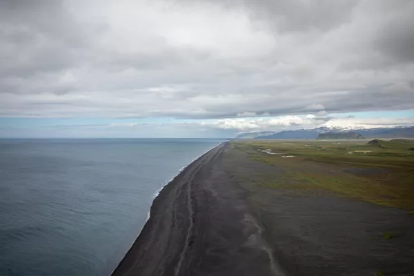 Playa de arena negra en la costa de Islandia vista desde lo alto, con el Atlántico gris, llanuras verdes y montañas nevadas