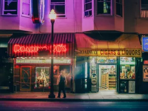 Fotografía de arte de Broadway en San Francisco de noche: neón rojo del Roaring 20’s, farola encendida y un transeúnte en movimiento