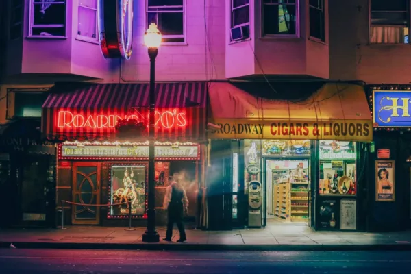 Fotografía de arte de Broadway en San Francisco de noche: neón rojo del Roaring 20’s, farola encendida y un transeúnte en movimiento